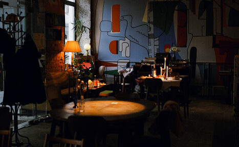 Арендовать Гастробар Дневник. Интерьер кафе с мебелью из дерева. Круглые и квадратные столы, стулья. На стенах абстрактные рисунки. Освещение мягкое, с настольными лампами и свечами. Пол темный, ковровое покрытие. Есть вешалки для одежды и комнатные растения.