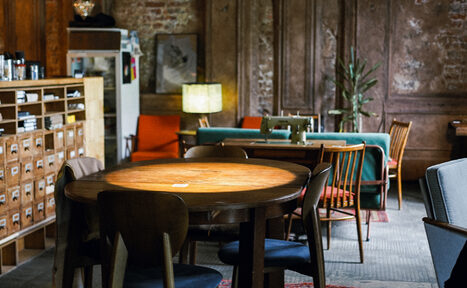 Арендовать Гастробар Дневник. Теплое, уютное пространство с круглым деревянным столом и стульями, вмещающее диваны и кресла. Стена с кирпичной отделкой, книжный стеллаж. Естественное освещение из лампы, ковры на полу. Оформлено в ретро-стиле с растениями.