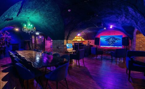 Арендовать GB Rooms(ГБ Румс). Лофт №1. Подвальное помещение с кирпичными стенами и арочными сводами. Длинный деревянный стол с мягкими стульями. На стене большой экран и компьютерный монитор. Фиолетовое и зеленое освещение, подвесная люстра. Деревянный пол, динамики по бокам экрана.