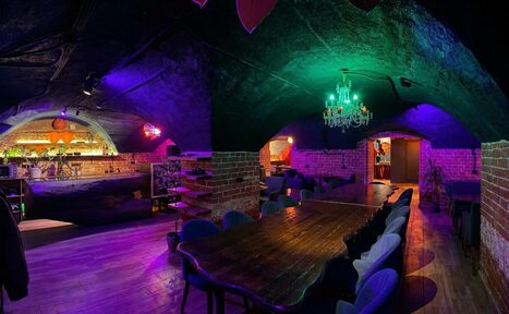 Арендовать GB Rooms(ГБ Румс). Лофт №1. Подвальное помещение с кирпичными стенами и арочным потолком. Длинный деревянный стол и мягкие стулья. Барная стойка в глубине. Цветное освещение: зелёные и фиолетовые оттенки. Люстра в центре. На полу деревянное покрытие.