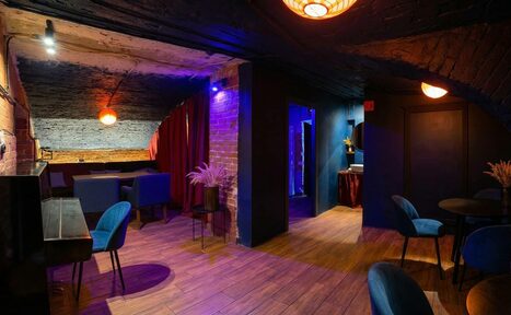 Арендовать GB Rooms(ГБ Румс). Лофт №1. Интерьер помещения: кирпичные стены, деревянный пол, черный и синий цвет стен, закрыто красной занавесью. Мебель: синие кресла, столы, пианино. Освещение: теплые светильники, потолочные лампы. Декор: вазы с растениями.