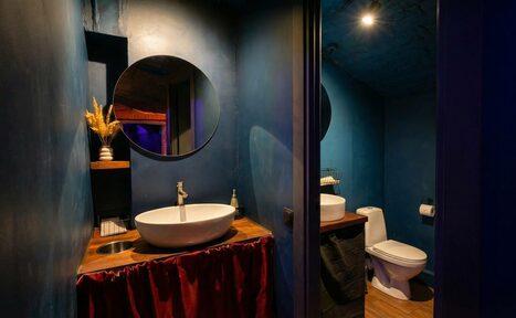 Арендовать GB Rooms(ГБ Румс). Лофт №1. Туалетная комната с темно-синими стенами, раковиной на деревянной тумбе с красной занавеской, круглым зеркалом, светильником, туалетом и декоративной вазой на полке. Освещение мягкое, материалы деревянные и текстильные.
