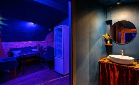 Арендовать GB Rooms(ГБ Румс). Лофт №1. Помещение с зоной отдыха и санузлом. В зоне отдыха диван, стол, стул, холодильник, кирпичная и синяя стена. В санузле круглое зеркало, раковина, лампы, синяя стена, красная тканевая юбка под раковиной. Залито синим освещением.