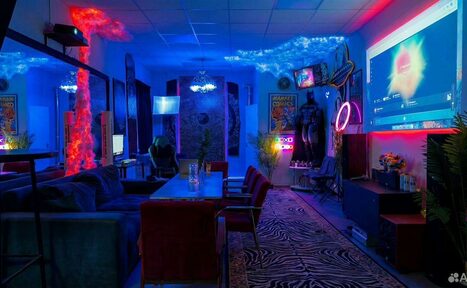 Арендовать GB Rooms(ГБ Румс). Лофт №2. Современная комната с диванами и креслами, столом, проектором на стене, разноцветной неоновой подсветкой, зеркалами, плакатами на стенах, растениями, декоративным освещением, акустической панелью и ковром с зебровым узором.