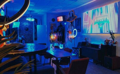 Арендовать GB Rooms(ГБ Румс). Лофт №2. Креативное пространство с неоновым освещением. В центре прямоугольный стол с стульями, рядом круглый стол со стульями. Неоновые знаки, проекция на стене. Декор в стиле комиксов, мягкая мебель, цветы в вазе. Игровая консоль на тумбе.