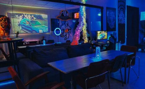 Арендовать GB Rooms(ГБ Румс). Лофт №2. Современное пространство с диванами и деревянным столом, окруженное стульями. На стенах арт-работы, синий и неоновый свет, два экрана с изображениями. Яркие световые акценты и декоративные элементы.