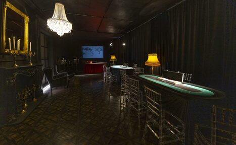 Арендовать Лофт Космос. Зал Президент. Игровое пространство с покерными столами, прозрачными стульями и классическими диванами. На стене проектор с изображением. Темные шторы, люстра и настольные лампы обеспечивают освещение. Паркетный пол, декоративные каминные элементы, зеркало и подсвечники.