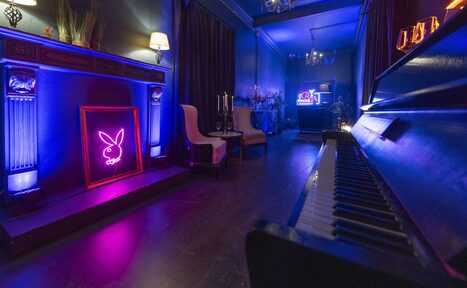 Арендовать Лофт Космос. Зал Президент. Стильное пространство с пианино и мягкими креслами, украшенное неоновыми знаками. Темные стены, шторы и деревянный пол. Встроенные светильники и настенные бра создают атмосферное освещение. Декор в виде подсвечников и зелени на камине.