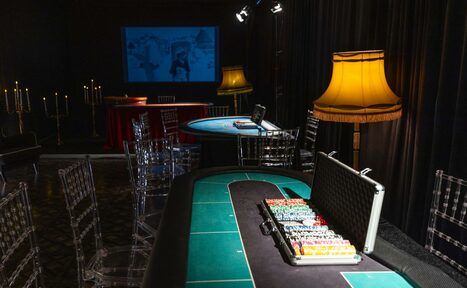 Арендовать Лофт Космос. Зал Президент. Комната с игорными столами, прозрачными стульями и черными занавесками. Настольные лампы с желтыми абажурами, кейс с фишками, проектор с экраном на стене, канделябры. Тёмное освещение, полы с узором.