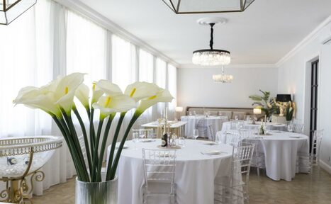 Арендовать Лофт Космос. Зал Президент. Просторный банкетный зал с круглыми столами, накрытыми белыми скатертями, прозрачными стульями, высокими окнами, люстрами и букетами искусственных цветов. Светлые стены, лампы на столах, светлый пол.