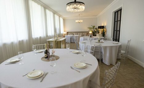 Арендовать Лофт Космос. Зал Президент. Большая, светлая комната с несколькими круглыми столами, накрытыми белыми скатертями. Прозрачные стулья, приборы, бокалы и тарелки на столах. Светильники с кристаллами. Белые стены и большие окна с полупрозрачными шторами. Зеленые растения в углу.