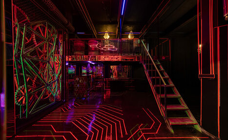 Арендовать Лофт Космос. Зал Ночной Клуб. Темное помещение в индустриальном стиле с неоновым освещением в красных и синих тонах. Лофт с металлической лестницей на второй ярус. Геометрические узоры на стенах и полу. Видна барная стойка и сетчатые конструкции.