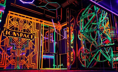 Арендовать Лофт Космос. Зал Ночной Клуб. Интерьер с неоновыми графиками и светящимися линиями на стенах и потолке. Присутствует слово "ONSPACE" на стене. Геометрические узоры и яркие цвета: оранжевый, зеленый, фиолетовый. Элементы декора напоминают шестерни.