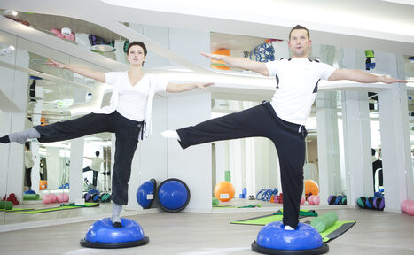 Арендовать Face & Body(Лицо и Тело). Спортивное помещение с зеркальными стенами и светлым деревянным полом. Два человека выполняют упражнения на синих балансировочных полусферах. На полу видны оборудование для фитнеса: гимнастические коврики, мячи и гантели. Яркое накладное освещение.