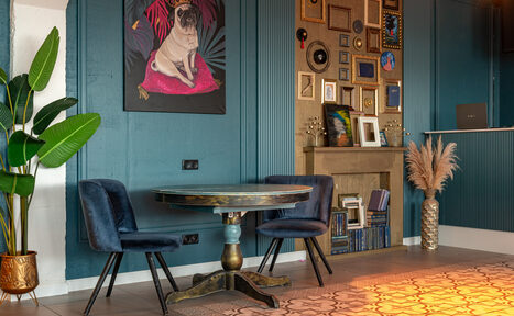 Арендовать Лофт Май. Стильное пространство с верандой. Пространство с синими стенами и декоративными панелями, стол и два синих стула, настенное искусство с изображением собаки, галерея из рамок, растение в горшке, декоративная каминная полка с книгами и аксессуарами, светлый пол с узорчатым ковром.