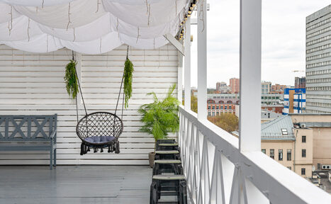 Арендовать Лофт Май. Стильное пространство с верандой. Терраса с деревянным полом и стенами, навесом из белого текстиля. Подвесное кресло и скамейка, вокруг несколько растений в горшках. Декоративные световые гирлянды. Вид на городские здания.