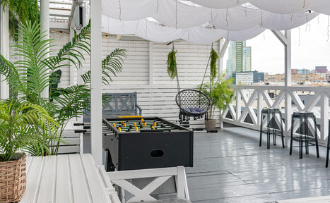 Арендовать Лофт Май. Стильное пространство с верандой. Терраса с белыми деревянными стенами и полом, оснащена настольным футболом, стульями из черного металла и плетеным креслом. Подвешены белые ткани, горшки с зелеными растениями. Панорамный вид на город.