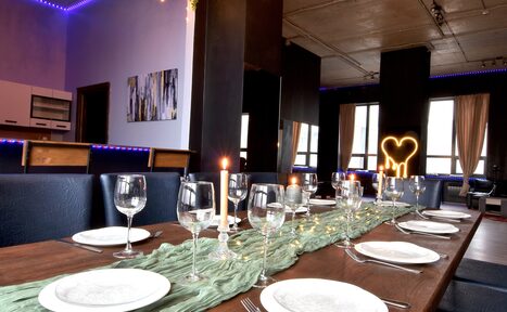 Арендовать Элегантный лофт с хорошим звуком и балконом. Просторное помещение с деревянным столом, сервированным бокалами и тарелками. На стене картина, рядом открытая кухня с мебелью. Потолок с накладным освещением, стены светлые. Светодиодная подсветка по периметру, большое окно со шторами.