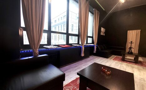 Арендовать Элегантный лофт с хорошим звуком и балконом. Лофт-пространство с черной кожаной мебелью, низким черным столом, большими окнами с бежевыми шторами, светлый пол, стены темного цвета, в углу — кальян, на стене картина, есть декоративная подсветка.