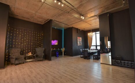 Арендовать Элегантный лофт с хорошим звуком и балконом. Просторное современное помещение с бетонным потолком и светлым деревянным полом. Чёрные стены с гирляндами, зона отдыха с двумя креслами, столик, обеденный стол со стульями. Большое окно с занавесью, зеркало, подсветка. Декорации на стенах.