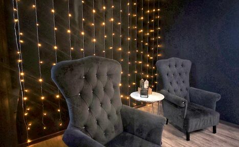 Арендовать Элегантный лофт с хорошим звуком и балконом. Два темных мягких кресла в комнате с деревянным полом. Между креслами стоит круглый белый стол с декором на поверхности. Темная стена задрапирована нитями теплых огоньков.