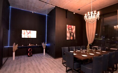 Арендовать Элегантный лофт с хорошим звуком и балконом. Темная комната с высоким потолком и бетонными элементами. В центре большой деревянный стол с синими стульями. На стене картина и телевизор. В углу динамики. Хрустальная люстра и светодиодное освещение. Серые полы, большие окна с занавесками.