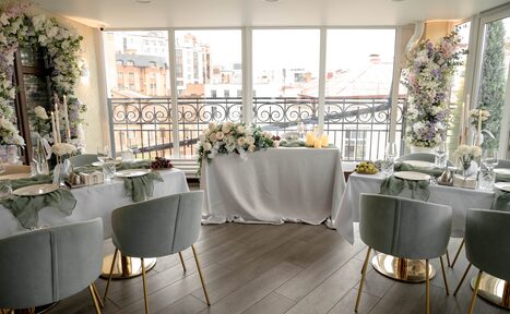 Арендовать Ресторан Небо Казани. Пространство с панорамными окнами, видом на город, три накрытых стола с белыми скатертями, декоративные свечи и цветочные композиции, серые мягкие стулья с золотыми ножками, пол из темного дерева, украшенные цветами стены.