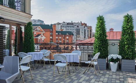 Арендовать Ресторан Небо Казани. Терраса с видом на городские здания, выложенная плиткой. Несколько круглых столов с белыми скатертями окружены складными стульями. Зелёные декоративные растения в горшках вдоль ограждения. Спокойный светлый день.