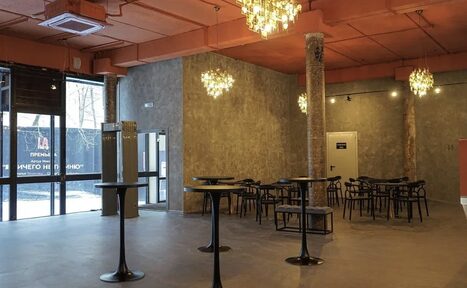 Арендовать Театр в Хамовниках. Просторное помещение с выполненными в индустриальном стиле стенами, равномерно расположенными столиками и стульями. Выделяются стеклянные двери, декоративные светильники и ярко оранжевый потолок с видимой вентиляцией.