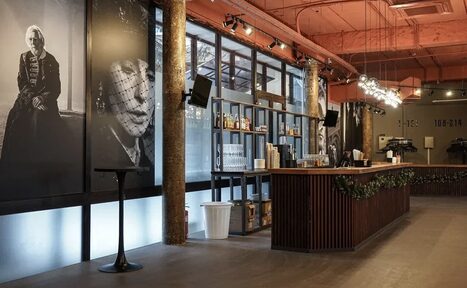 Арендовать Театр в Хамовниках. Барное пространство в индустриальном стиле с бетонными колоннами и деревянной стойкой. Большие чёрно-белые портреты украшают стены. На потолке открытые коммуникации. Освещение представлено подвесными лампами. Полки с бутылками, мусорная корзина.