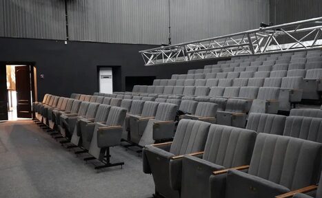 Арендовать Театр в Хамовниках. Аудиторный зал с рядной расстановкой серых кресел с подлокотниками. Пол покрыт ковролином, стены темные. Над креслами металлическая конструкция для освещения. Справа от двери виден шкаф. Освещение из дверного проема.