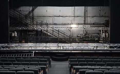 Арендовать Театр в Хамовниках. Аудитория со сценой и рядами темных кресел. На сцене установлена металлическая конструкция. Задняя стена с серыми панелями и трубами. Освещение тусклое, фон серый.