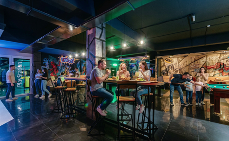 Арендовать Площадка для праздников Spark. Пространство включает зону с высокими столами и стульями, бильярдный стол. Яркое освещение, темные полы. На стенах яркие граффити, изображены персонажи. Надпись "PlayStation". Композитный материал мебели, яркие цвета в интерьере.