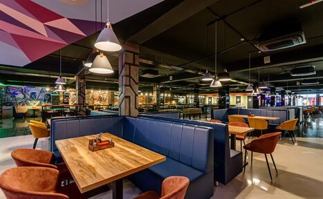 Арендовать Площадка для праздников Spark. Просторное помещение с деревянными столами и синими диванами-кабинами. Коричневые стулья с мягкой обивкой. Современные подвесные светильники. Пол из светлого материала. Стены с граффити. Открытые коммуникации на потолке.