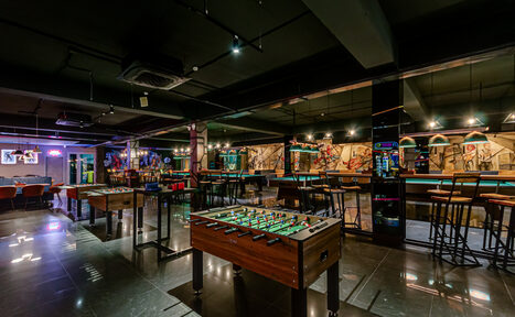 Арендовать Площадка для праздников Spark. Современное помещение для досуга с настольным футболом, барной стойкой и стульями. Интерьер выполнен в тёмных тонах, на стенах - абстрактный арт. Пол блестящий, освещение точечное, подвесное. Видна вывеска неоновая и телевизор на стене.