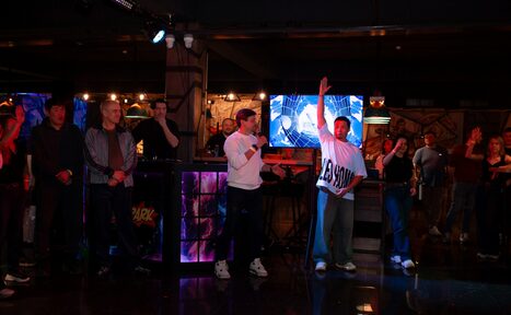 Арендовать Площадка для праздников Spark. Изображение интерьера с темным освещением, искусственные лампы создают разноцветное освещение. На переднем плане группа людей, один человек держит микрофон. На стене экран с яркими изображениями. Пол отражает свет.