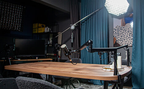 Арендовать Студия для записи подкастов "Искусство Подкаста". Подкаст-студия с деревянным столом, на котором установлены микрофоны на стойках. Стулья вокруг стола, световое оборудование. Фон оформлен шторами. Присутствует монитор, черные полки с техникой и коробками. Геометрический постер на стене.