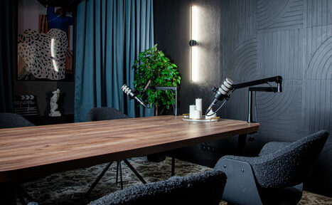 Арендовать Студия для записи подкастов "Искусство Подкаста". Комната с длинным деревянным столом и тремя темными креслами. На столе два микрофона на стойках. Справа вертикальный светильник на стене. На заднем плане синие шторы, зелёное растение и стены с черным рельефным узором.