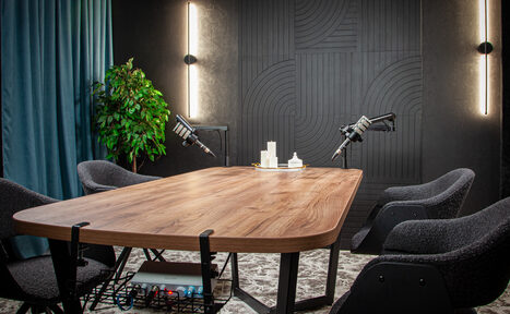 Арендовать Студия для записи подкастов "Искусство Подкаста". Современное студийное пространство с деревянным столом, четырьмя серыми креслами и микрофонами на стойках. Освещение вертикальными светильниками, тёмные стеновые панели с геометрическим узором, большое растение у стены. Ковровое покрытие приглушенных тонов.