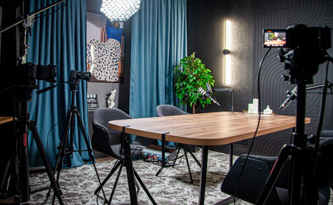 Арендовать Студия для записи подкастов "Искусство Подкаста". Современное студийное пространство с деревянным столом и серыми креслами. Съемочное оборудование, включая камеры и свет. Темные стены, голубые шторы, зеленое растение. Вертикальный светильник на стене. Настенный постер и акустическая панель.