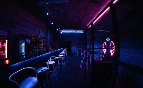 Арендовать PVZ CLUB (ПВЗ Клуб). Барная зона с длинной стойкой и высокими стульями. Кирпичные стены, неоновые огни с оттенками синего и розового. На стене неоновая фигура крыльев с ореолом. Холодильник и полки с напитками. Пол тёмный, потолок индустриального стиля.