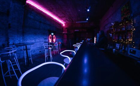 Арендовать PVZ CLUB (ПВЗ Клуб). Клубное пространство с длинной стойкой и высокими стульями, кирпичные стены, неоновая подсветка фиолетового и синего цвета, полки с напитками, таблички с меню на стене, приглушенное освещение.