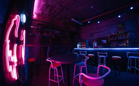 Арендовать PVZ CLUB (ПВЗ Клуб). Интерьер барного пространства с неоновым освещением. Круглый стол и стулья в центре, барная стойка с высокими стульями справа. На стене неоновая вывеска. Цвета: розовый, синий. Потолок и стены из кирпича, пол из дерева. Светодиодные лампы на стенах.