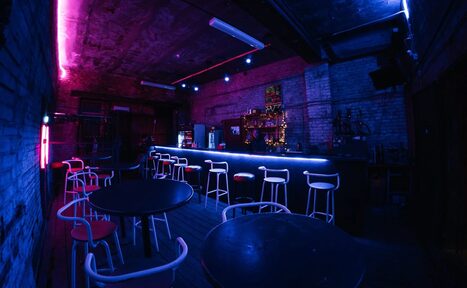 Арендовать PVZ CLUB (ПВЗ Клуб). Барное помещение с кирпичными стенами, темным деревянным полом и неоновым голубым и розовым освещением. Барная стойка с высокими стульями, круглые черные столы с белыми стульями, светильники на потолке.