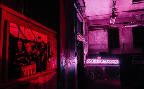 Арендовать PVZ CLUB (ПВЗ Клуб). Тёмное пространство с неоном, барная стойка с вывеской "DISCO", стены с граффити, потолок с обшарпанной краской, тусклое освещение красного оттенка, на стенах плакаты, в том числе с изображениями людей, провода на потолке.