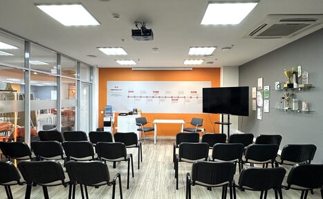 Арендовать Конференц-зал. Конференц-зал с рядами черных стульев, белыми и оранжевыми стенами. На переднем плане телевизор на штативе, белый стол, стеклянные двери. Светлое деревянное покрытие пола, потолочные светильники, проектор. На стенах полки с наградами и плакатами.