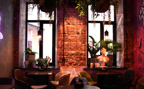 Арендовать Винный бар “Дело не в вине”. Лофт-пространство с кирпичными стенами. Два окна с чёрными рамами, декоративные растения на подоконниках и бюст. Низкие чёрные столы, разноцветные стулья. Подвесное освещение, неоновые элементы, стены с граффити. Атмосферное освещение.