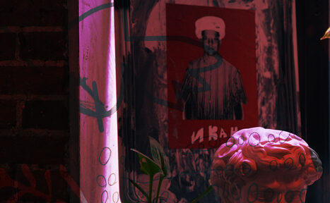 Арендовать Винный бар “Дело не в вине”. Арт-пространство с кирпичной стеной, на которой висит постер с изображением человека в шефском колпаке. На переднем плане бюст розового цвета. Освещение фиолетовое и розовое, частично изображён зелёный лист растения.