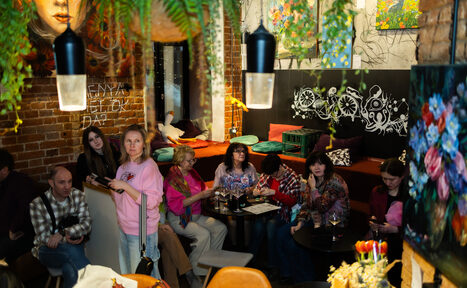 Арендовать Винный бар “Дело не в вине”. Кафе или лаунж пространство с кирпичными стенами и настенными рисунками. Мягкие сиденья с подушками, столики, люди сидят и стоят, растения свисают с потолка. Освещение с подвесными лампами, картины на стенах.