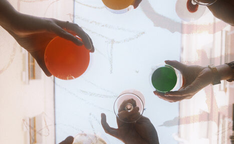 Арендовать Винный бар “Дело не в вине”. Руки людей держат разноцветные стеклянные бокалы на фоне светлого неба и размытых очертаний зданий.